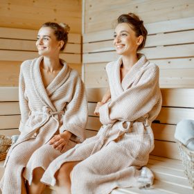 Two young beautiful girlfriends in bathrobes sitting together while relaxing in the luxury sauna